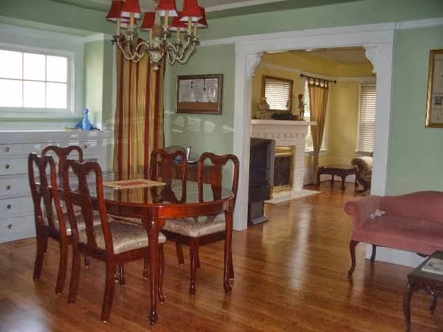 A dining room with a polished wooden dining table and six matching chairs with cushioned seats. The room has light green walls and a chandelier with red lampshades hanging above the table. There is a window with striped curtains and a white dresser beneath it. Through an open doorway, a living room with a fireplace, yellow walls, and wooden flooring is visible. The living room has a sofa and a small table near the window with blinds.