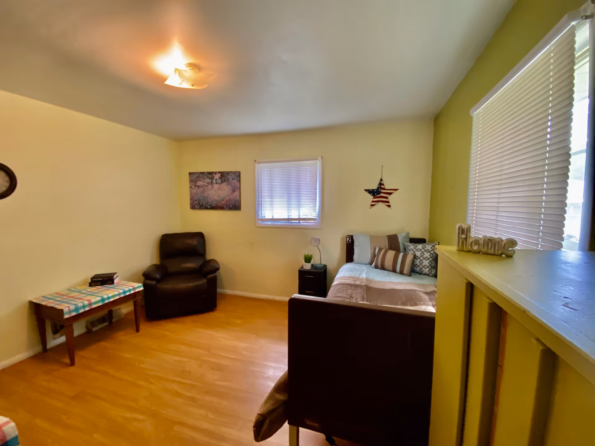 A small, cozy bedroom with light yellow walls and a wooden floor. The room contains a single bed with multiple pillows and a beige blanket, a small black nightstand with a lamp and a small plant, a black recliner chair, and a wooden table covered with a colorful checkered cloth. There are two windows with white blinds, a wall decoration of an American flag star, and a painting on the wall. A decorative 'Home' sign is placed on a yellow partition near the window.