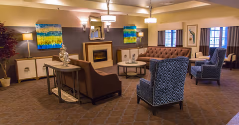 A cozy living room with a brown tufted sofa, two blue patterned armchairs, a brown loveseat, and a round coffee table in the center. The room features a fireplace with a mirror above it, two colorful abstract paintings on the wall, and several lamps providing warm lighting. There are windows with curtains on the right side and a small table with decorative items near the loveseat.