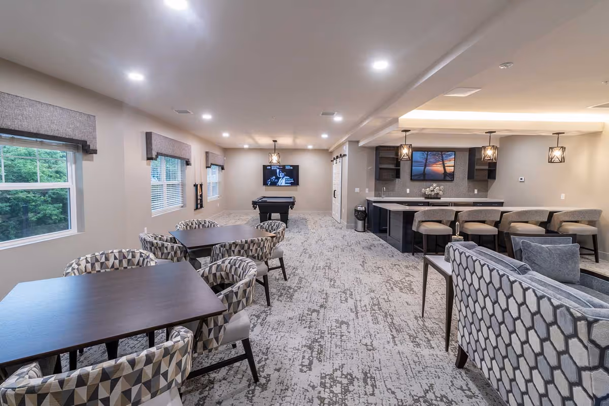 A spacious senior living common area with multiple seating options including patterned armchairs around dark wood tables, a gray patterned sofa, and a bar counter with cushioned stools. The room features large windows with gray valances, recessed ceiling lights, pendant lights above the bar, a wall-mounted TV, and a pool table in the background.
