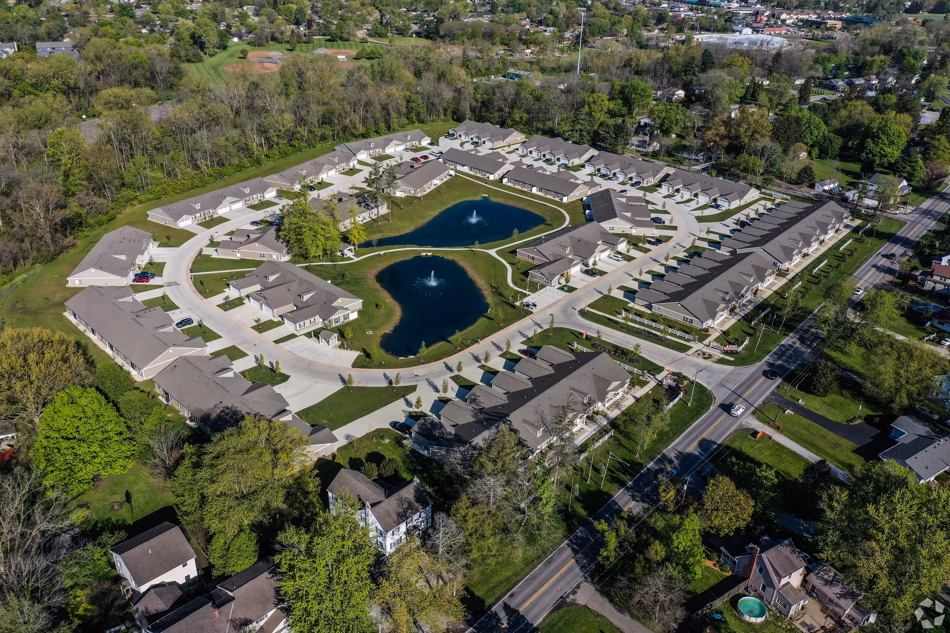 Aerial view of a residential community with multiple single-story buildings arranged around two small ponds with fountains. The area is surrounded by trees and greenery, with roads and parking spaces visible. The neighborhood includes a mix of houses and townhomes, with a main road running alongside the community.