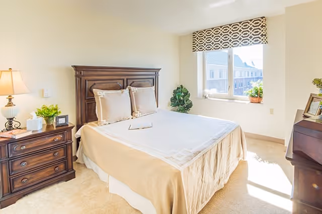 Sunlit bedroom with a neatly made bed, wooden headboard, nightstand with lamp, and a window with a patterned valance.