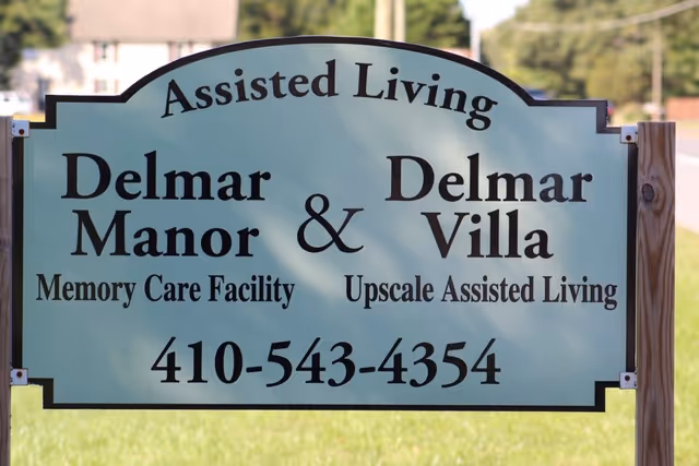 A freestanding sign for Delmar Manor & Delmar Villa Assisted Living and Memory Care displaying the facility name and phone number.
