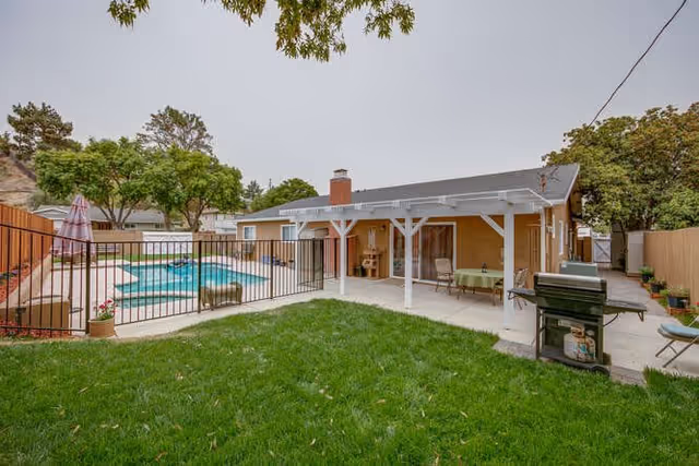 Outdoor area of a senior living facility featuring a fenced swimming pool with a patio umbrella, a covered patio with seating and a table, a barbecue grill, and a well-maintained grassy lawn surrounded by trees and fencing.