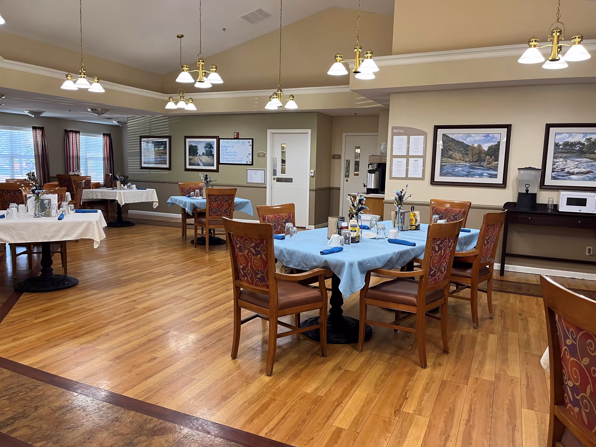 A dining room in a senior living facility with several tables covered in blue and white tablecloths, each set with glasses, napkins, and small flower arrangements. The room has wooden floors, multiple hanging light fixtures, framed landscape paintings on the walls, and windows with curtains letting in natural light.
