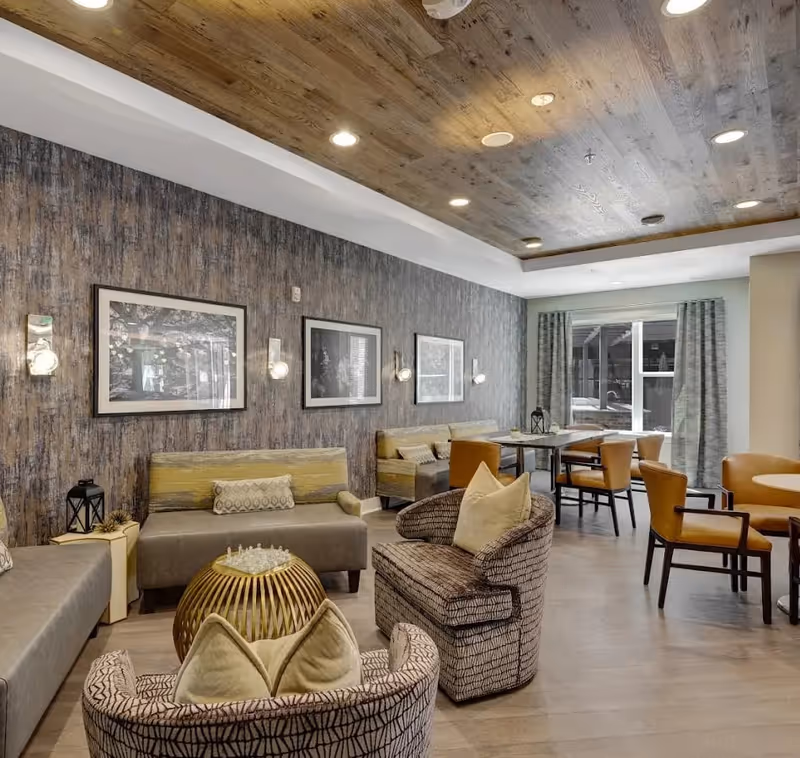 A cozy senior living facility common area with patterned armchairs and cushioned benches arranged around a small round table with a chess set. The room features a wood-paneled ceiling with recessed lighting, textured wallpaper with framed black and white photos, and large windows with curtains letting in natural light. Additional seating includes a dining table with chairs in the background.