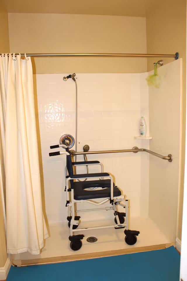 Tiled shower stall with a wheeled shower chair/commode, grab bars, showerhead, curtain and a bottle of soap on a shelf.