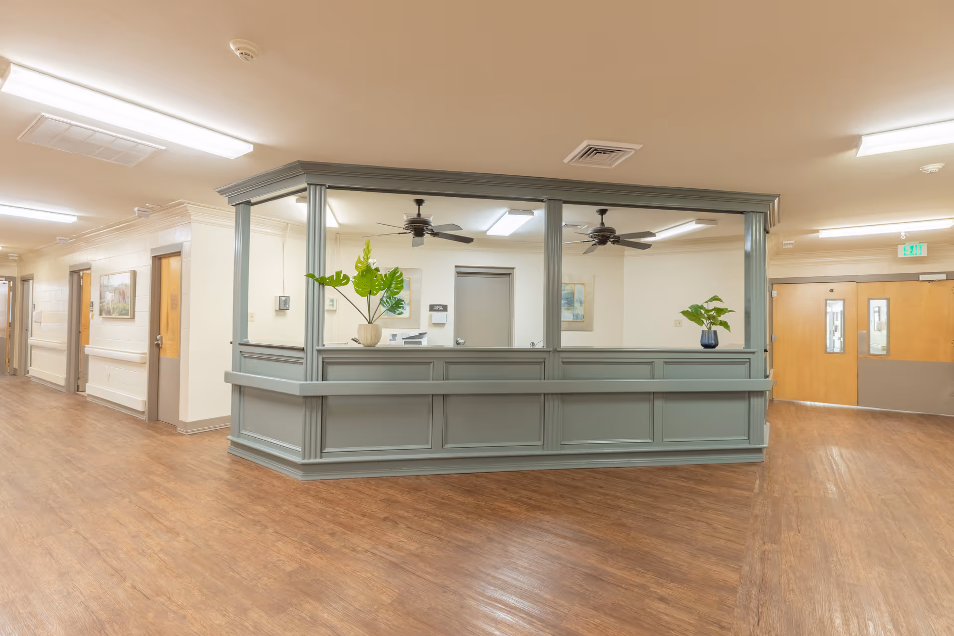 Interior view of a senior living facility reception area with a large gray reception desk in the center. The desk has two potted plants on it and ceiling fans above. The floor is wood, and there are hallways and doors visible in the background with fluorescent lighting overhead.