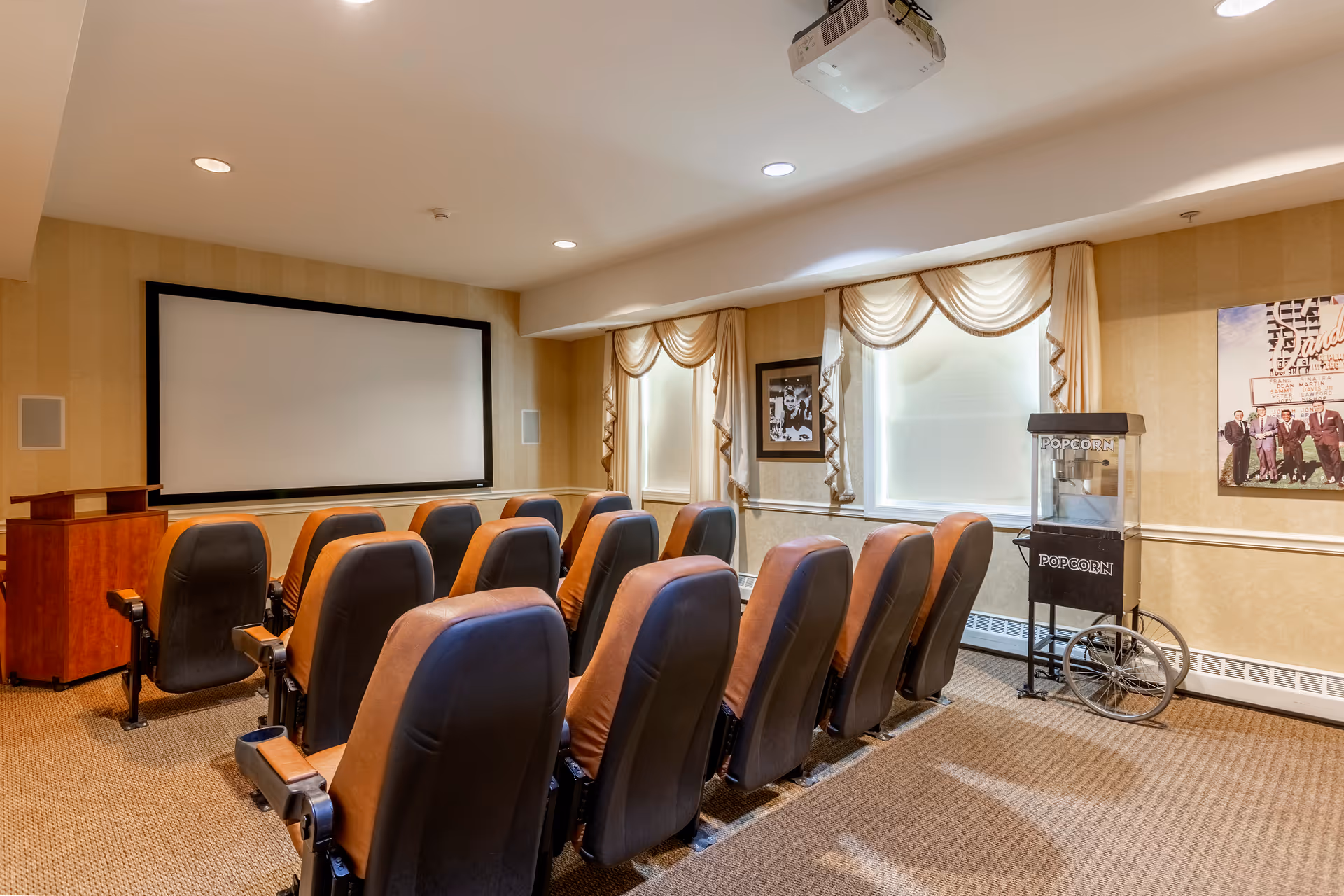 A small theater room with two rows of brown and black cushioned chairs facing a large blank projector screen. The room has beige walls with framed pictures, two windows with cream-colored curtains, a popcorn machine on the right side, and a ceiling-mounted projector.