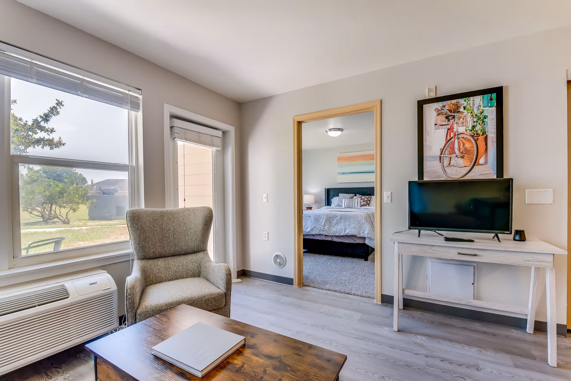 A bright living room area with a large window and a glass door letting in natural light. There is a comfortable armchair next to a wooden coffee table with a book on it. Across from the chair, a small white desk holds a flat-screen TV and a framed picture of a red bicycle. Through an open doorway, a bedroom with a bed and a colorful wall painting is visible.