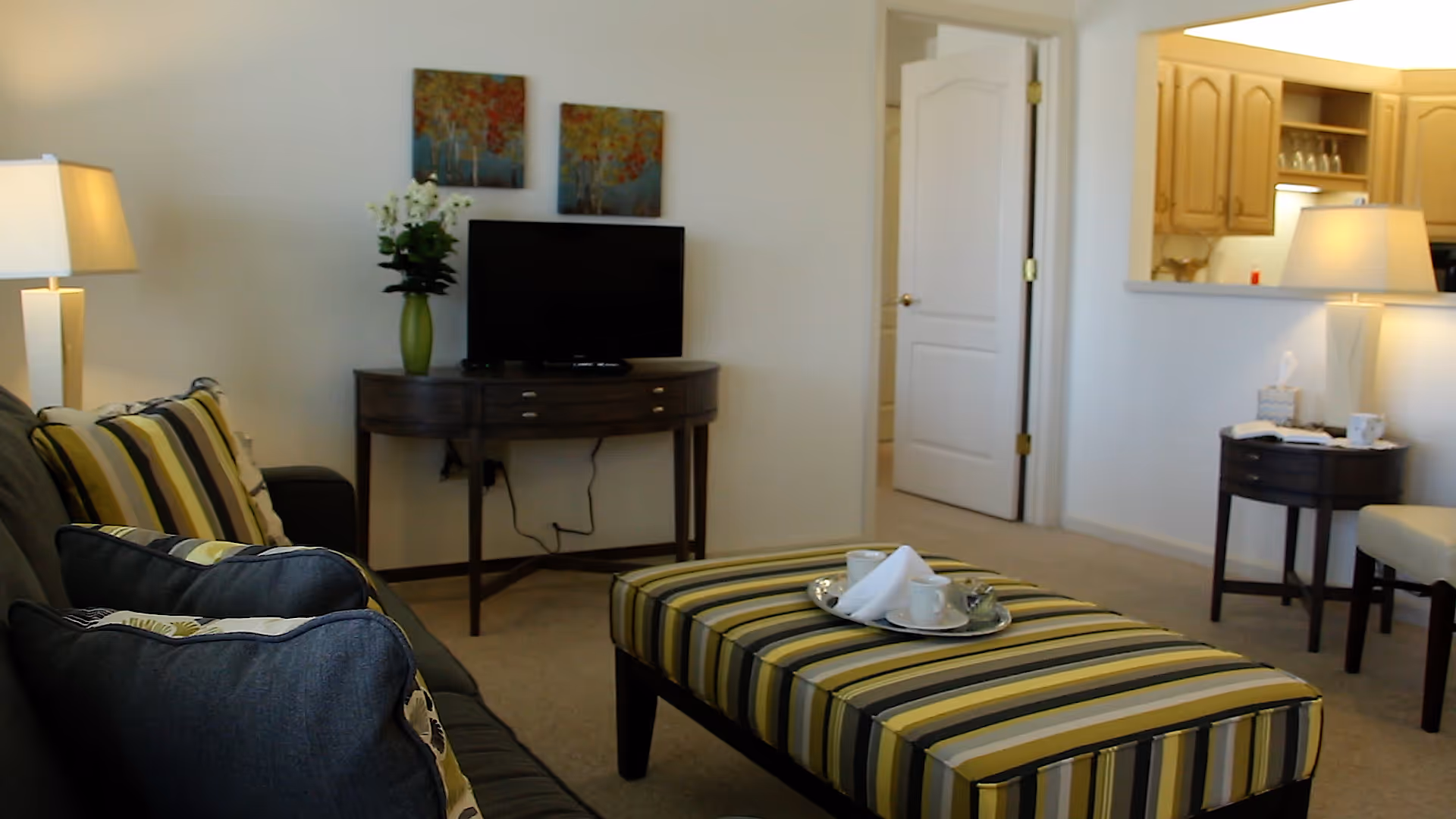 A cozy living room with a striped ottoman in the center, a dark sofa with multiple cushions on the left, a small wooden table with a flat-screen TV and a green vase with white flowers against the wall, two colorful paintings above the TV, a doorway leading to another room, and a kitchen area visible through a pass-through window on the right. Two table lamps provide warm lighting.