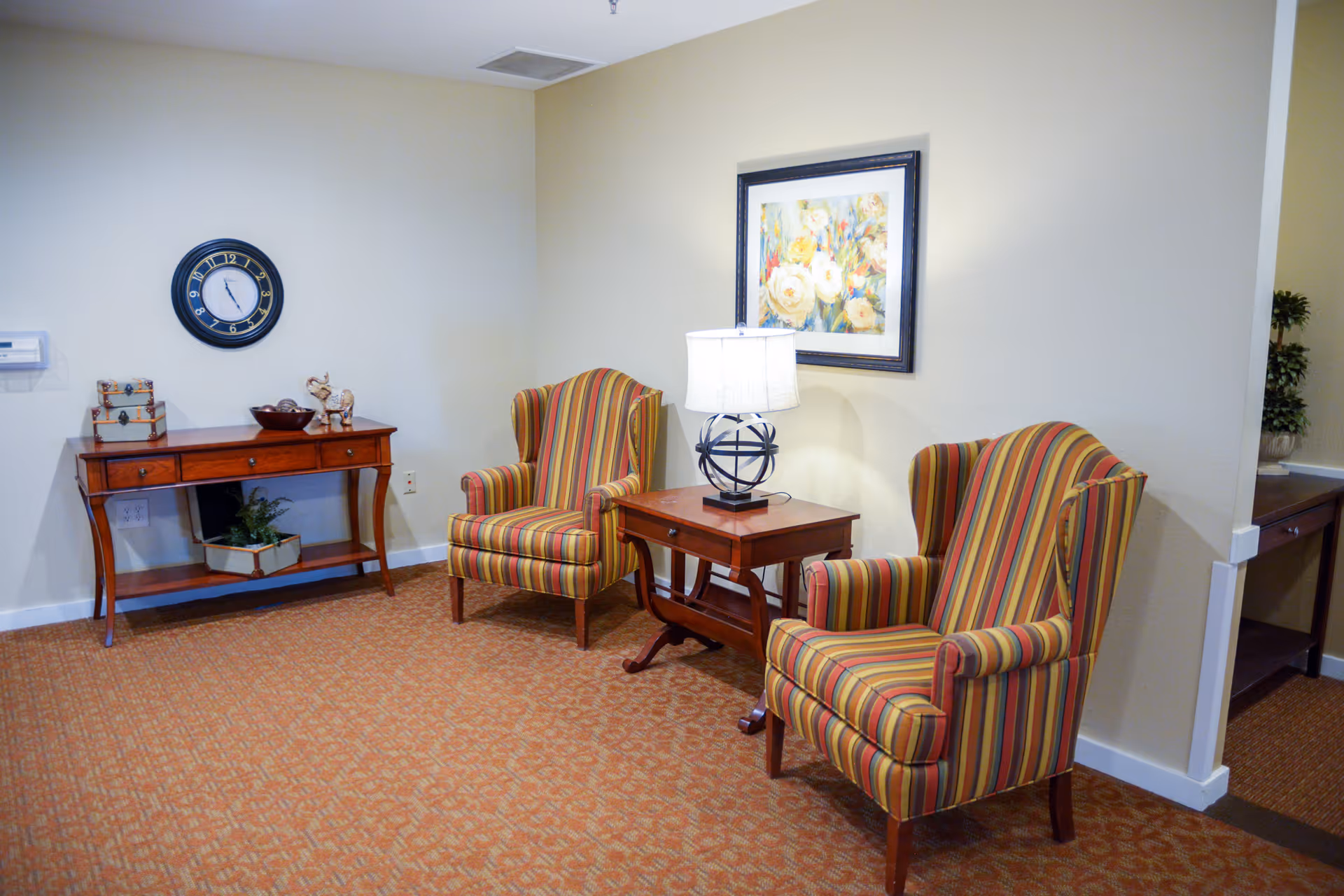 A cozy sitting area with two striped armchairs facing each other, a wooden side table with a decorative lamp between them, a wooden console table against the wall with decorative items, a round wall clock, and a framed floral painting on the wall.