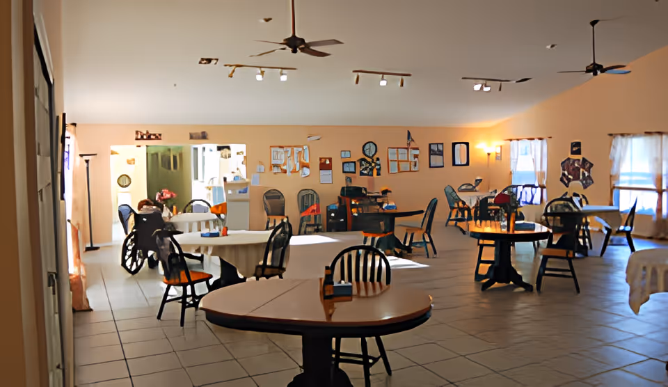 A spacious dining room with multiple round and rectangular tables surrounded by chairs. The room has peach-colored walls with bulletin boards and decorations, ceiling fans, and large windows with sheer curtains allowing natural light to enter. There is a wheelchair visible near one of the tables and a floor lamp in the corner.