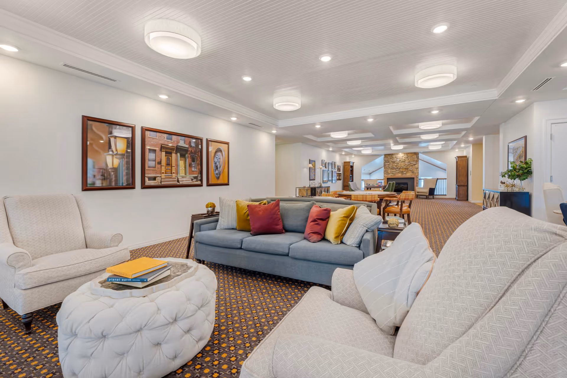 A spacious and well-lit living room area in a senior living facility featuring comfortable seating including a blue sofa with colorful pillows, two beige armchairs, and a white tufted ottoman with books on top. The room has patterned carpet, framed artwork on the walls, recessed lighting, and a stone fireplace in the background.