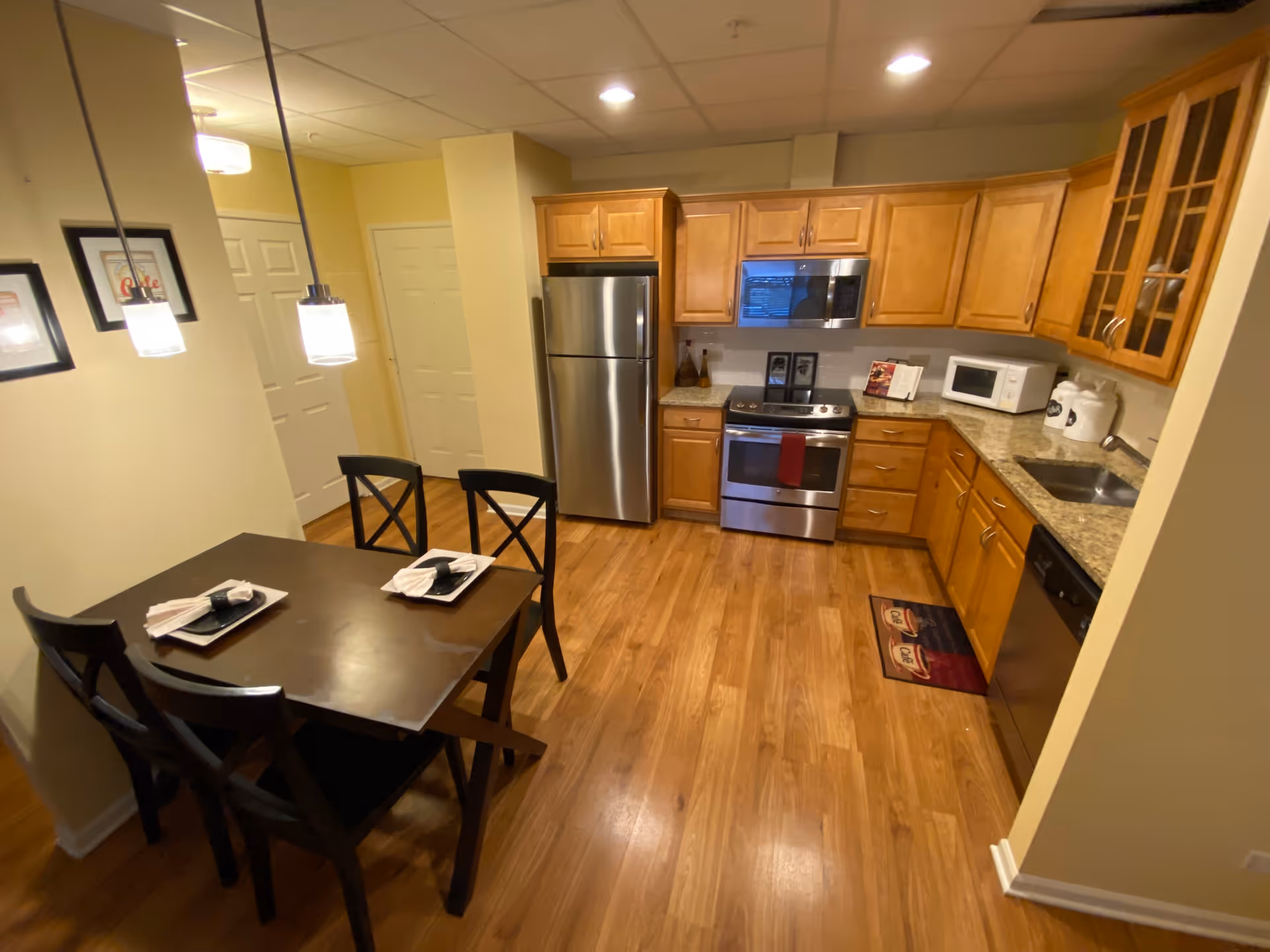 A kitchen and dining area with wooden cabinets, stainless steel refrigerator, stove, microwave, dishwasher, and a small sink. There is a dark wooden dining table with four chairs, set with plates and napkins. The floor is wooden, and two pendant lights hang above the dining table. The walls are painted light yellow.