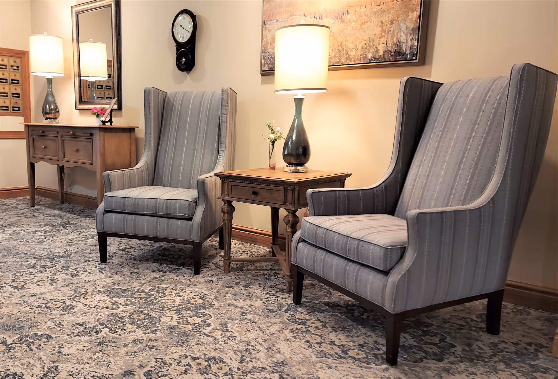 A cozy sitting area with two gray striped wingback chairs separated by a wooden side table with a lamp and small flower vase. Behind the chairs is a wall with a framed painting, a clock, and a wooden console table with another lamp and a mirror above it. The floor is covered with a patterned carpet.