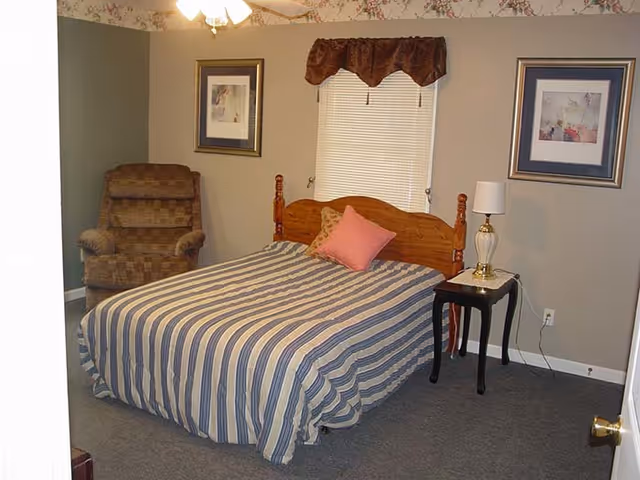 A furnished bedroom with striped bedspread, nightstand, lamp, chair, and window blinds.