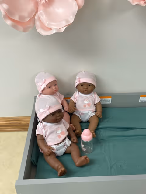 Three baby dolls in pink outfits and hats sit on a padded changing table next to a baby bottle beneath wall decorations.