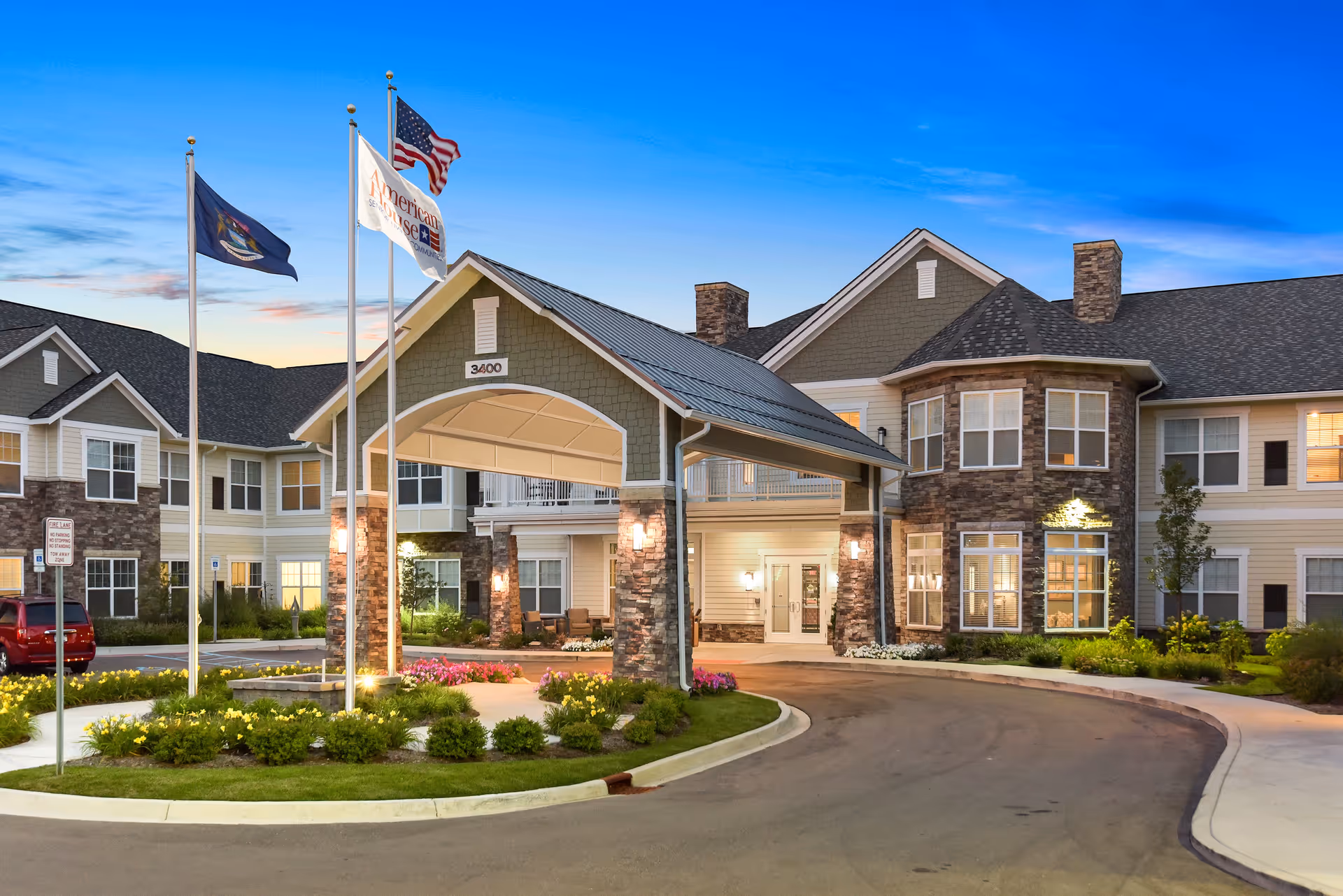 Exterior view of the American House Somerset senior living facility at dusk, showing a large covered entrance with stone pillars, well-maintained landscaping, and three flagpoles with flags flying. The building has multiple windows and a combination of stone and siding on the facade.
