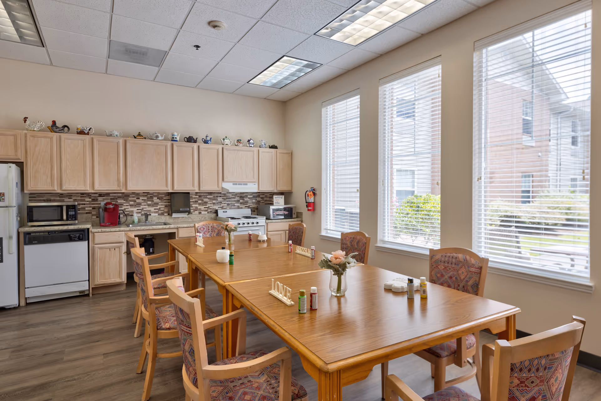 A bright kitchen and dining area with wooden cabinets, a refrigerator, microwave, dishwasher, stove, and a large wooden dining table surrounded by chairs with patterned upholstery. The table has small decorative items and flowers. Large windows with blinds let in natural light, and the floor is wood.