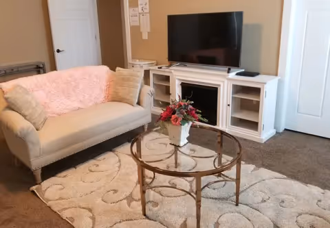 A cozy living room with a beige loveseat draped with a pink throw blanket and two beige pillows. In front of the loveseat is a round glass coffee table with a floral arrangement in a white vase. The room has a patterned beige carpet and a white entertainment unit with a flat-screen TV on top. There are two closed white doors visible in the background.