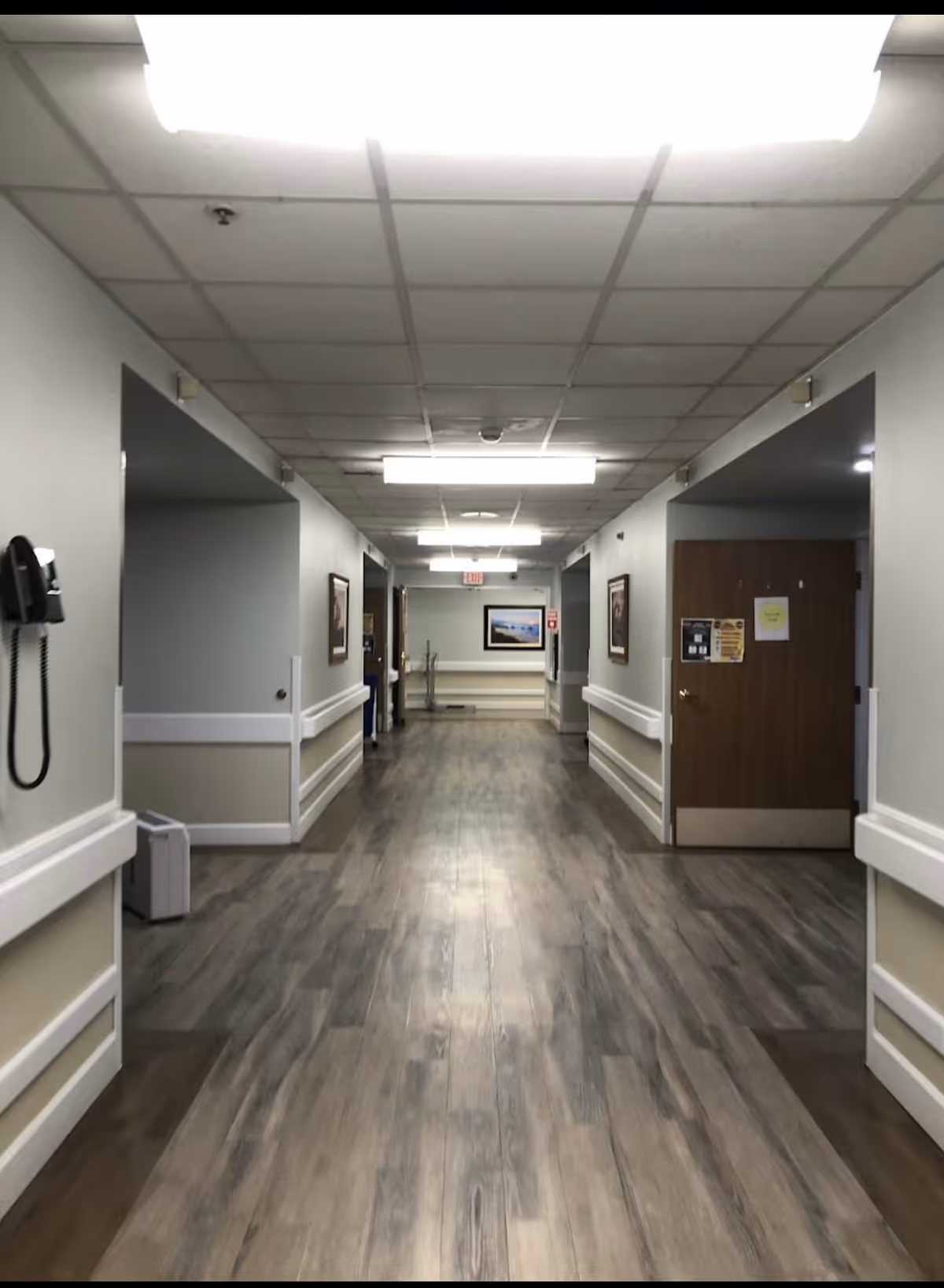 A long, empty hallway in a healthcare facility with wood-patterned flooring, white walls with beige lower panels, handrails on both sides, ceiling lights, a wall-mounted telephone on the left, framed pictures on the walls, and several doorways along the corridor.