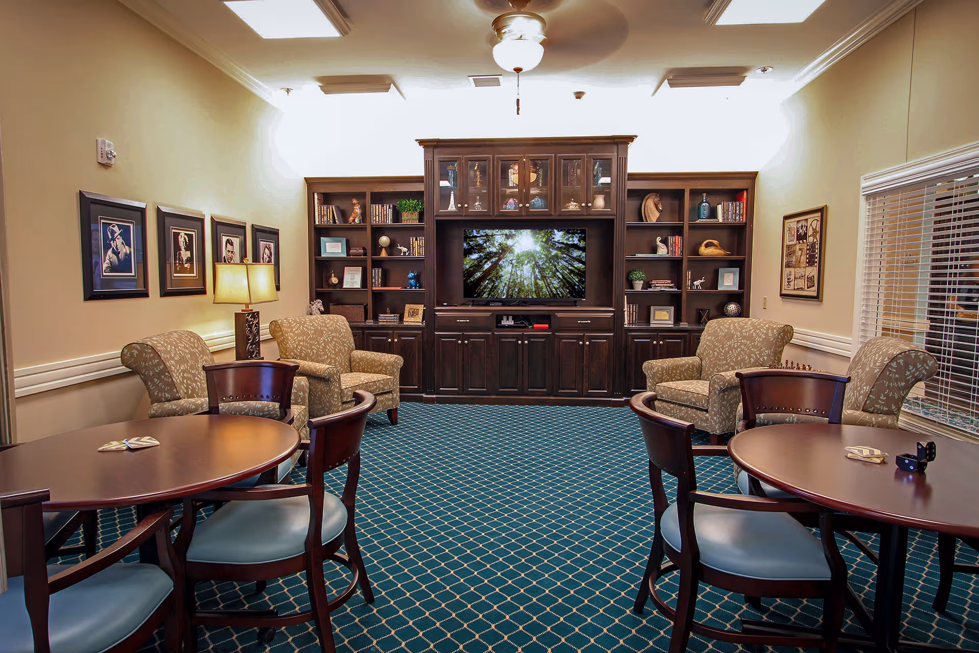 A cozy living room area with patterned blue carpet, two round wooden tables with chairs, four upholstered armchairs, a large dark wood entertainment center with a flat-screen TV displaying a forest scene, bookshelves with decorative items, framed pictures on the walls, a table lamp, and a ceiling fan with light.