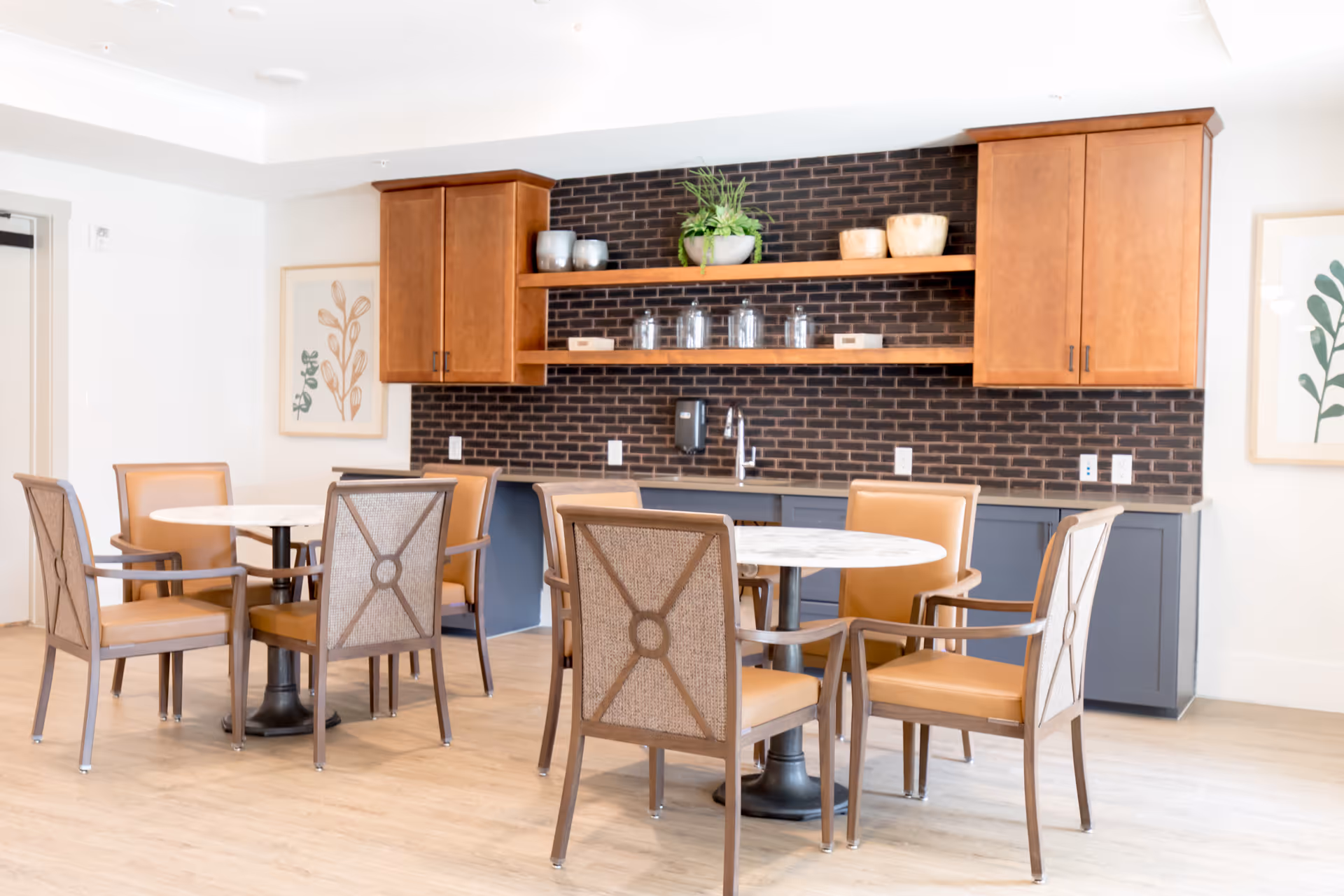 A bright dining area with two round tables surrounded by six chairs with tan cushions and woven backs. The background features a dark brick backsplash, wooden upper cabinets, open shelves with decorative items, and lower gray cabinets with a countertop and a sink.