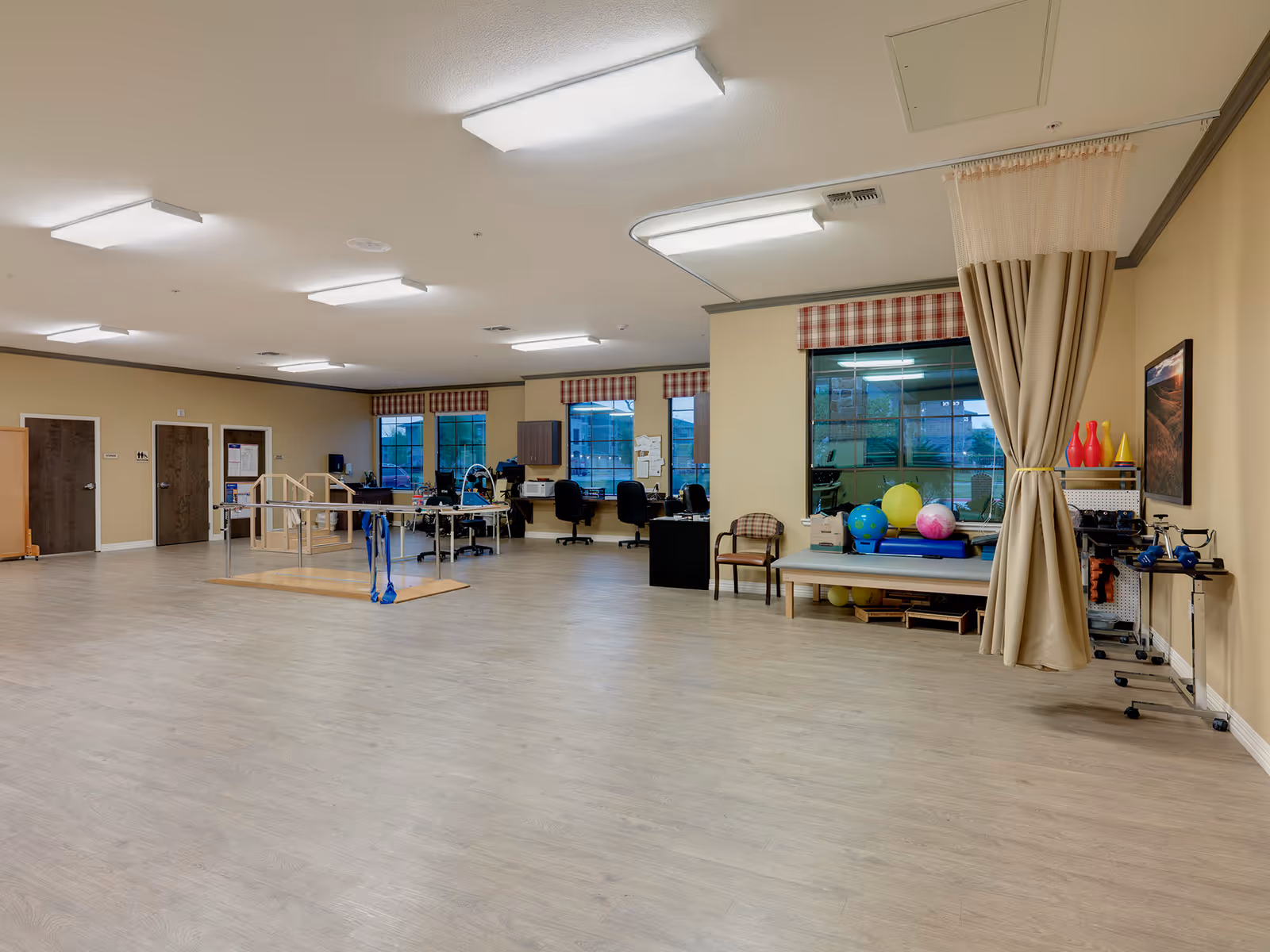 A spacious rehabilitation or therapy room with light wood flooring and beige walls. The room contains parallel bars for walking exercises, exercise balls, therapy equipment, chairs, desks with computers, and large windows with red and white checkered valances. A beige curtain is tied back on the right side of the room.