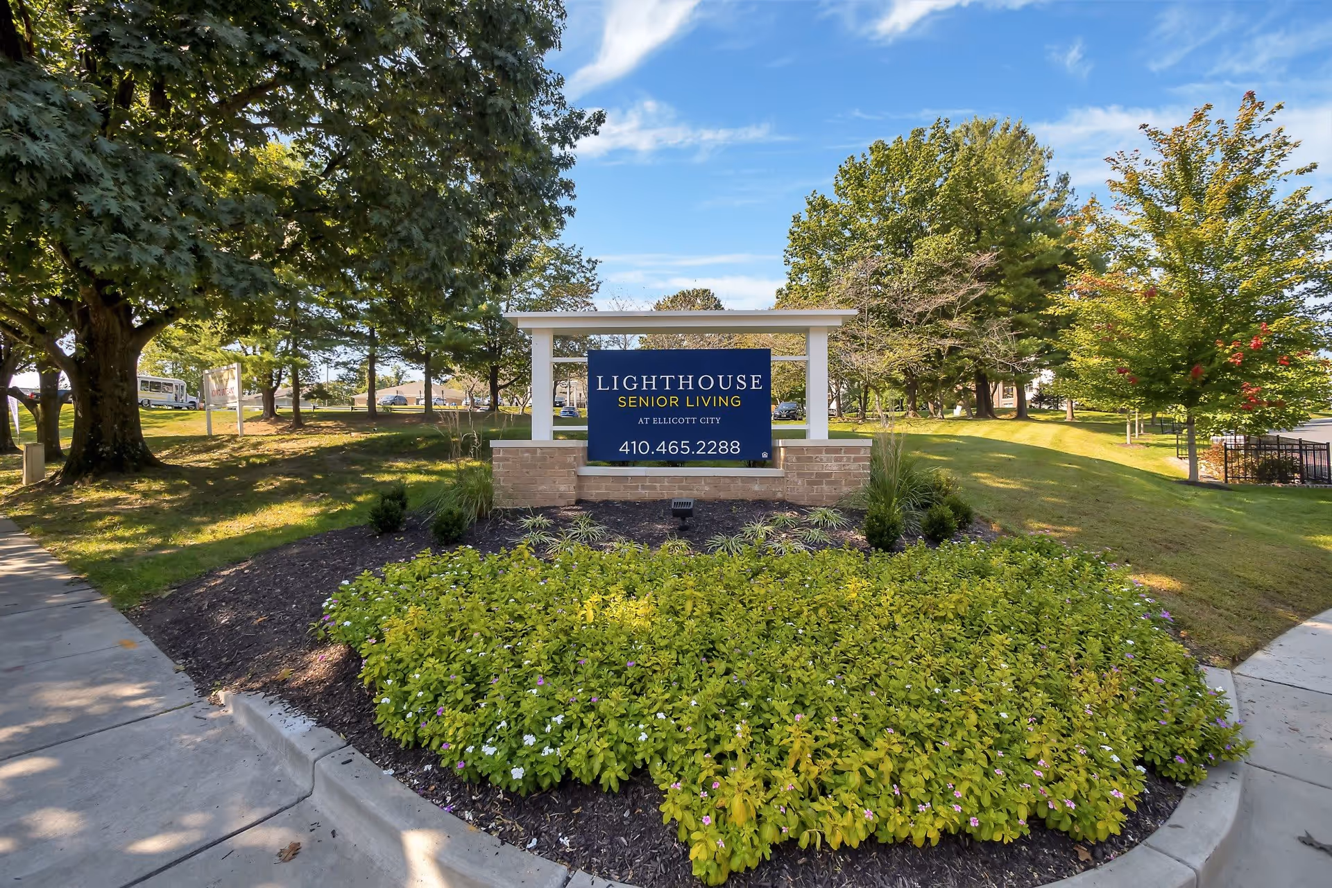 Outdoor view of the entrance sign for Lighthouse Senior Living at Ellicott City, surrounded by green trees, bushes, and a well-maintained garden bed under a blue sky with some clouds.