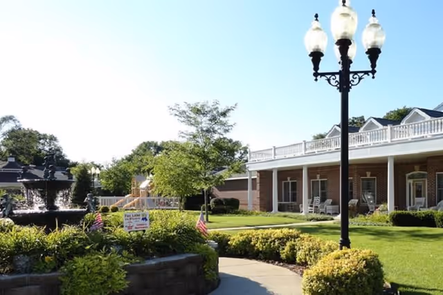 Outdoor view of Cottages of Fox Lake facility showing a landscaped garden with a fountain, green shrubs, a tree, American flags, a paved walkway, a black street lamp with three lights, and a brick building with a covered porch and white railings in the background.