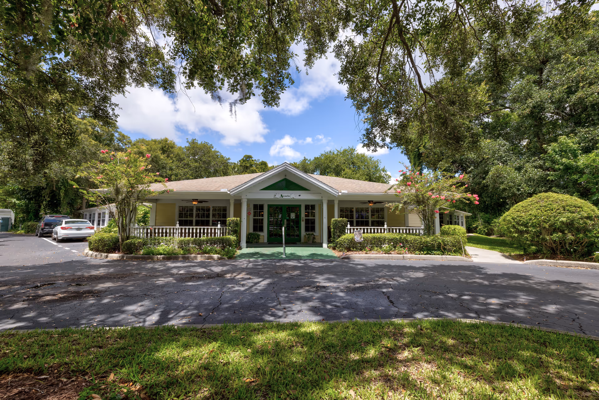 Front exterior view of a single-story building with a green entrance door, surrounded by trees and bushes under a partly cloudy sky. There are cars parked on the left side and a paved driveway in front.