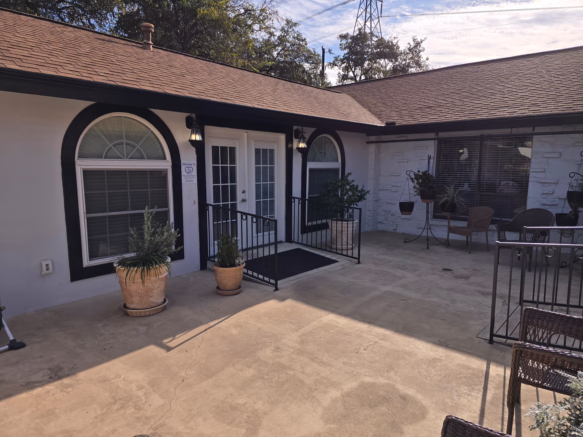 Outdoor patio area at Pipestone Place featuring a concrete floor, two large arched windows with black trim, a double glass door entrance with black railings and potted plants on either side. There are outdoor chairs and hanging plants on the right side under a roof overhang.
