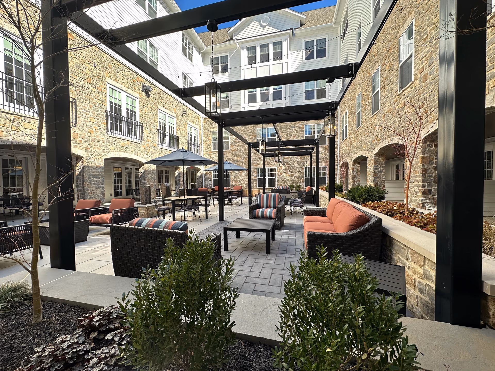 Outdoor courtyard area at Village Crossing at Worman's Mill featuring stone walls, multiple seating arrangements with cushioned chairs and sofas, tables with umbrellas, black metal pergolas with hanging lanterns, and some greenery in the foreground.