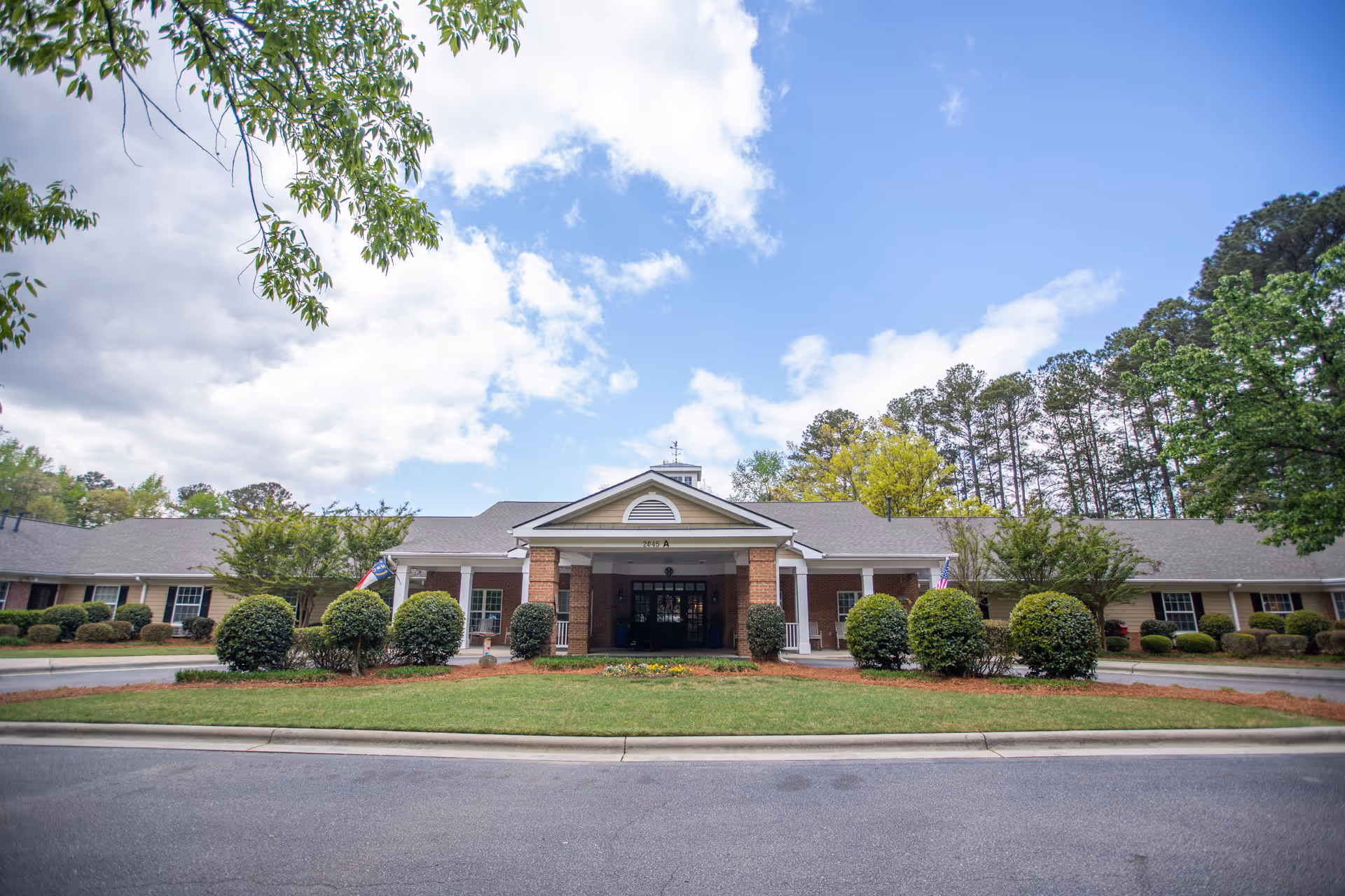 Front exterior view of a single-story senior living facility building with a covered entrance, surrounded by neatly trimmed bushes and trees under a partly cloudy sky.