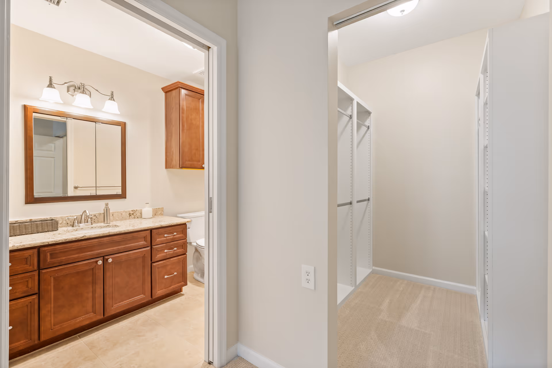 View of a bathroom with wooden cabinets, a granite countertop with a sink, a mirror with three lights above it, and a toilet partially visible. Adjacent to the bathroom is a walk-in closet with white shelving and beige carpet.