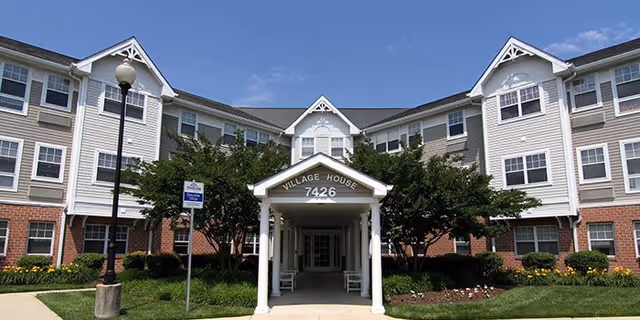 Front exterior of Village House Apartments showing a covered entrance portico labeled 7426 with landscaped grounds and a three-story white and brick facade.