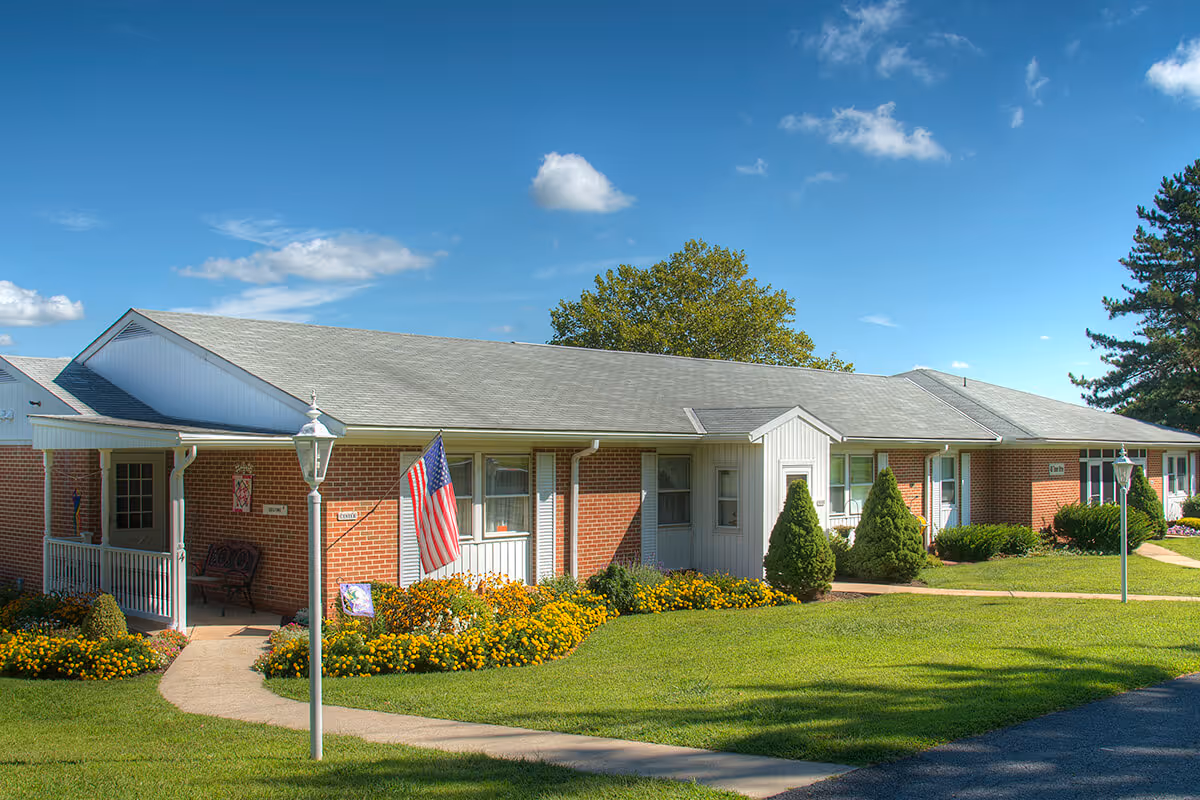 Single-story brick senior living building with a front porch, American flag, flower beds and a paved walkway under a blue sky.