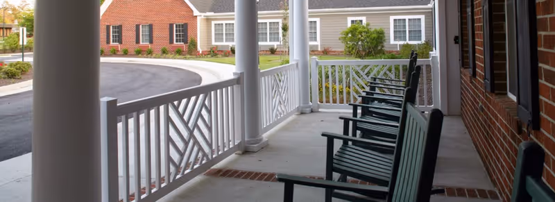 A covered porch area with several dark green rocking chairs lined up facing outward. The porch has white railings with a geometric pattern and white columns. In the background, there is a curved driveway and a building with brick and beige siding, windows, and some landscaping with bushes and flowers.