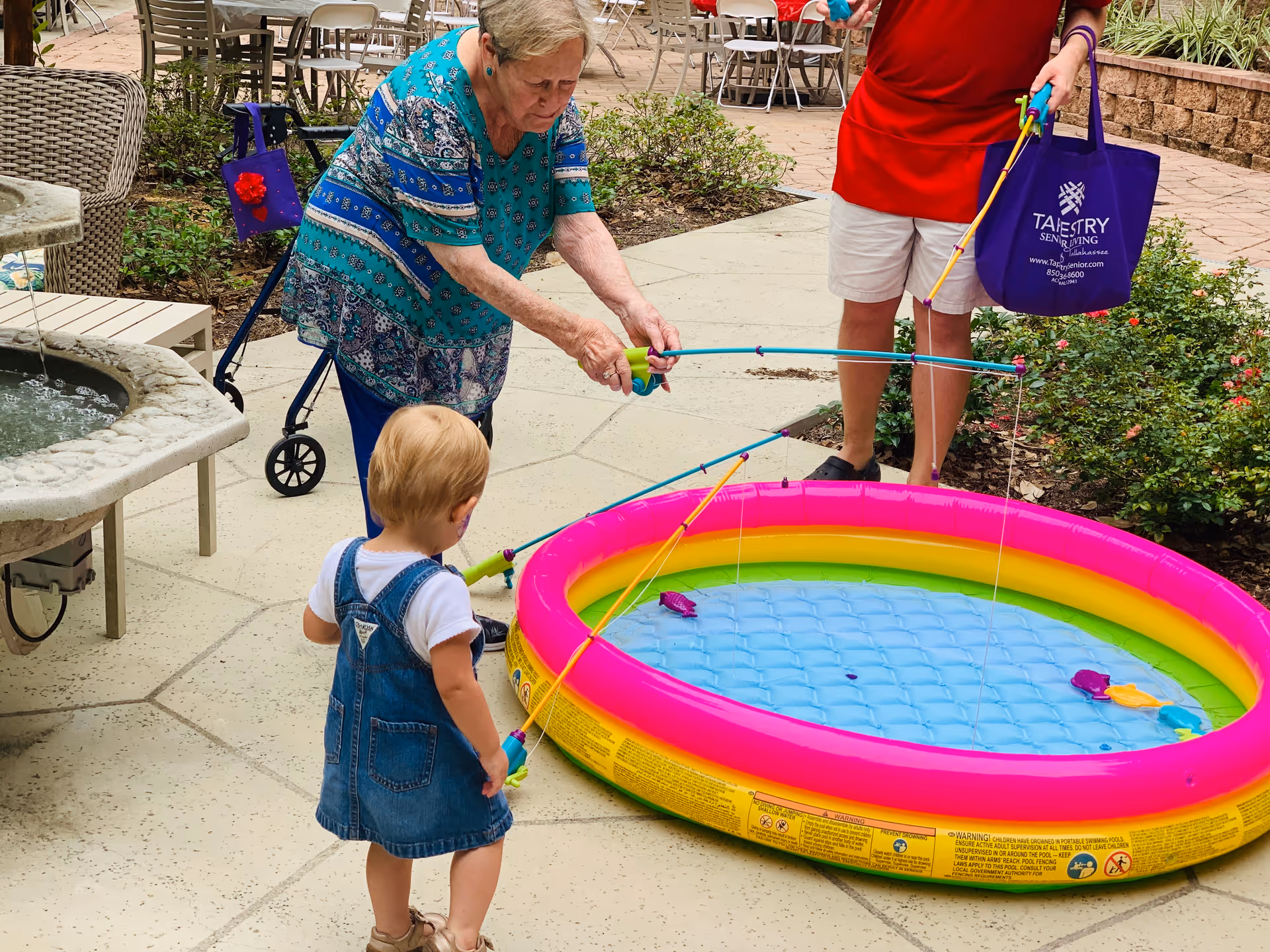 An elderly woman and a young child are playing with toy fishing rods at a small inflatable pool filled with water outdoors. Another person in a red shirt and white shorts is holding a purple tote bag with the Tapestry Senior Living Lakeshore logo. The setting is a paved patio area with garden plants and outdoor furniture.