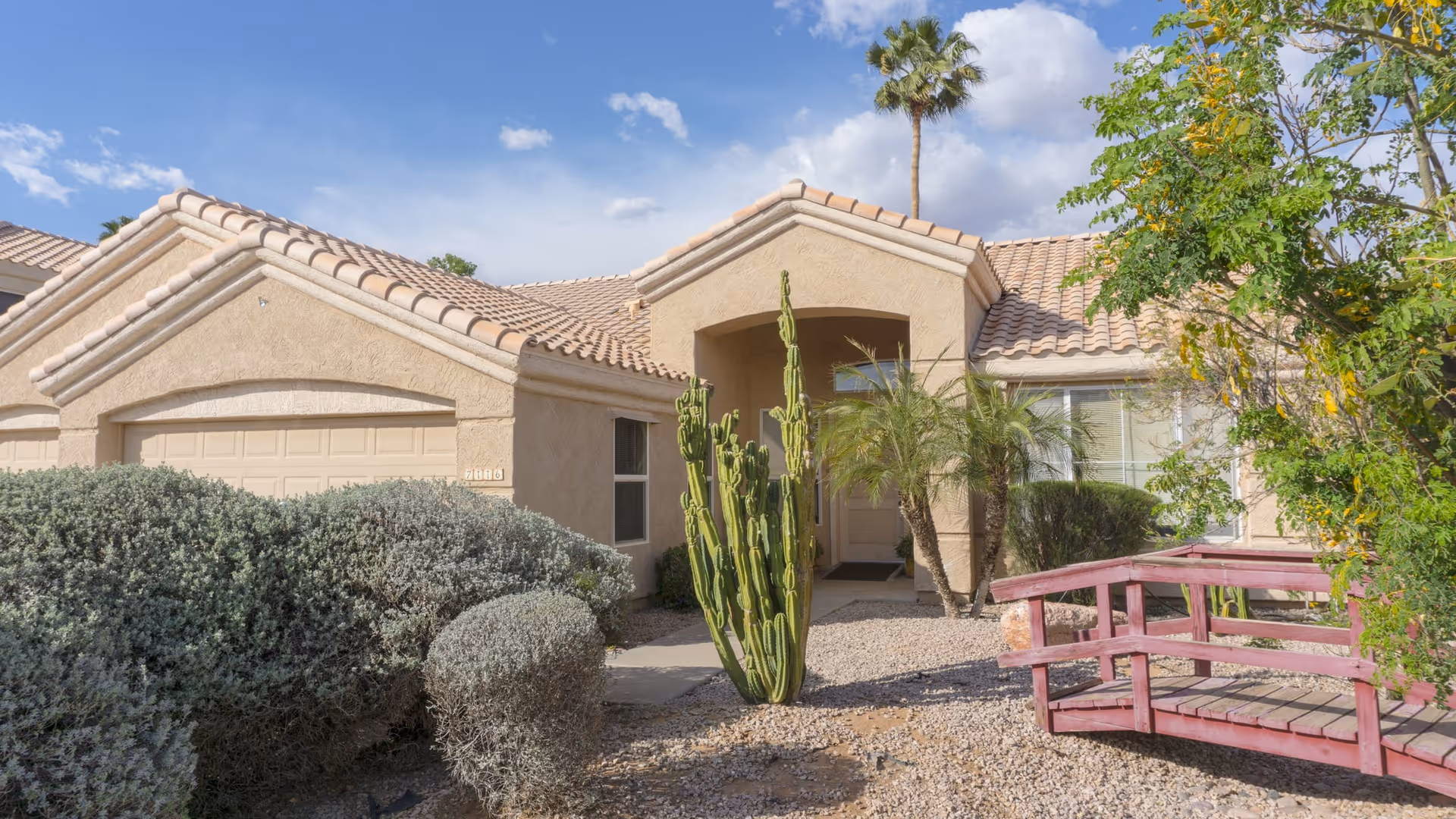 Exterior view of a single-story assisted living home with beige stucco walls and a tiled roof. The front entrance is surrounded by desert landscaping including cacti, bushes, and a small red wooden bridge over a dry gravel area. A tall palm tree is visible in the background under a partly cloudy blue sky.