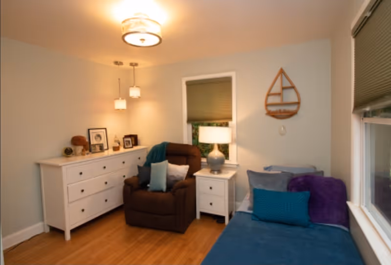 A cozy room with light-colored walls and wooden flooring featuring a brown recliner chair with multiple cushions, a white dresser with framed photos and decorative items, a white nightstand with a lamp, and a blue daybed with pillows and a purple throw blanket. Two windows with blinds and a wooden wall decoration shaped like a sailboat are also visible.