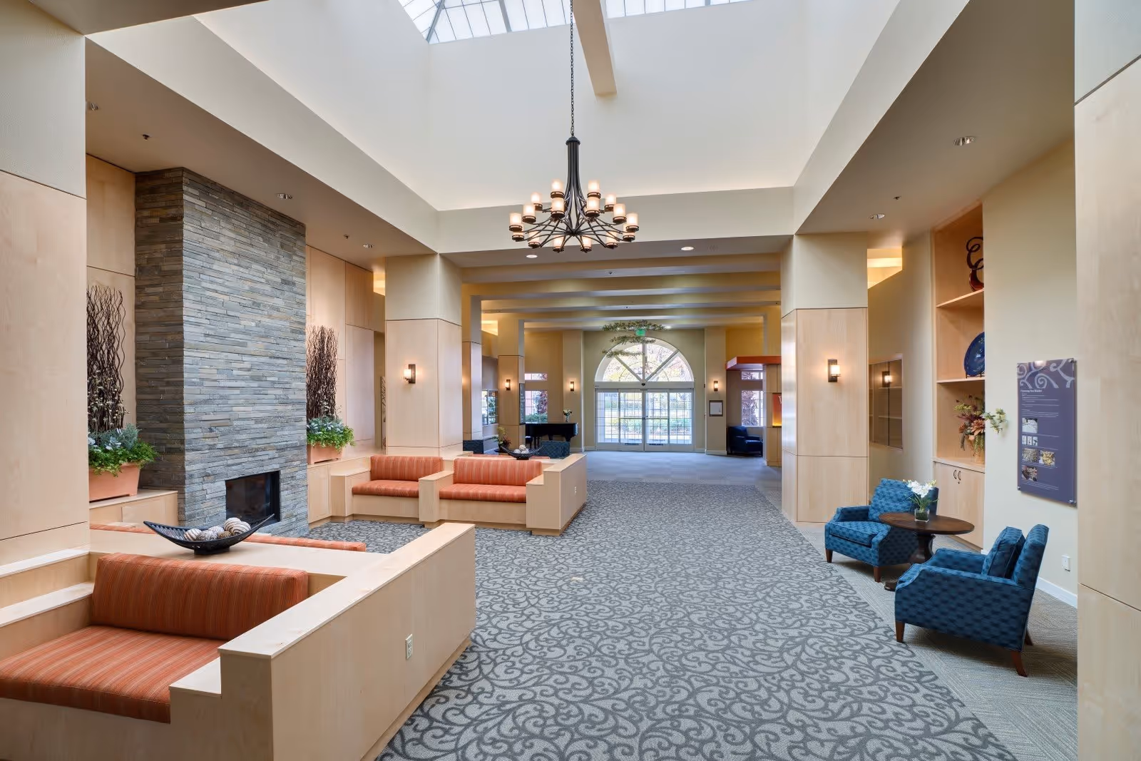 A spacious and well-lit common area with a high ceiling and large skylight. The room features a stone fireplace, several seating areas with orange and blue upholstered chairs, decorative plants, and a chandelier hanging from the ceiling. Large windows and glass doors are visible at the far end, allowing natural light to enter.