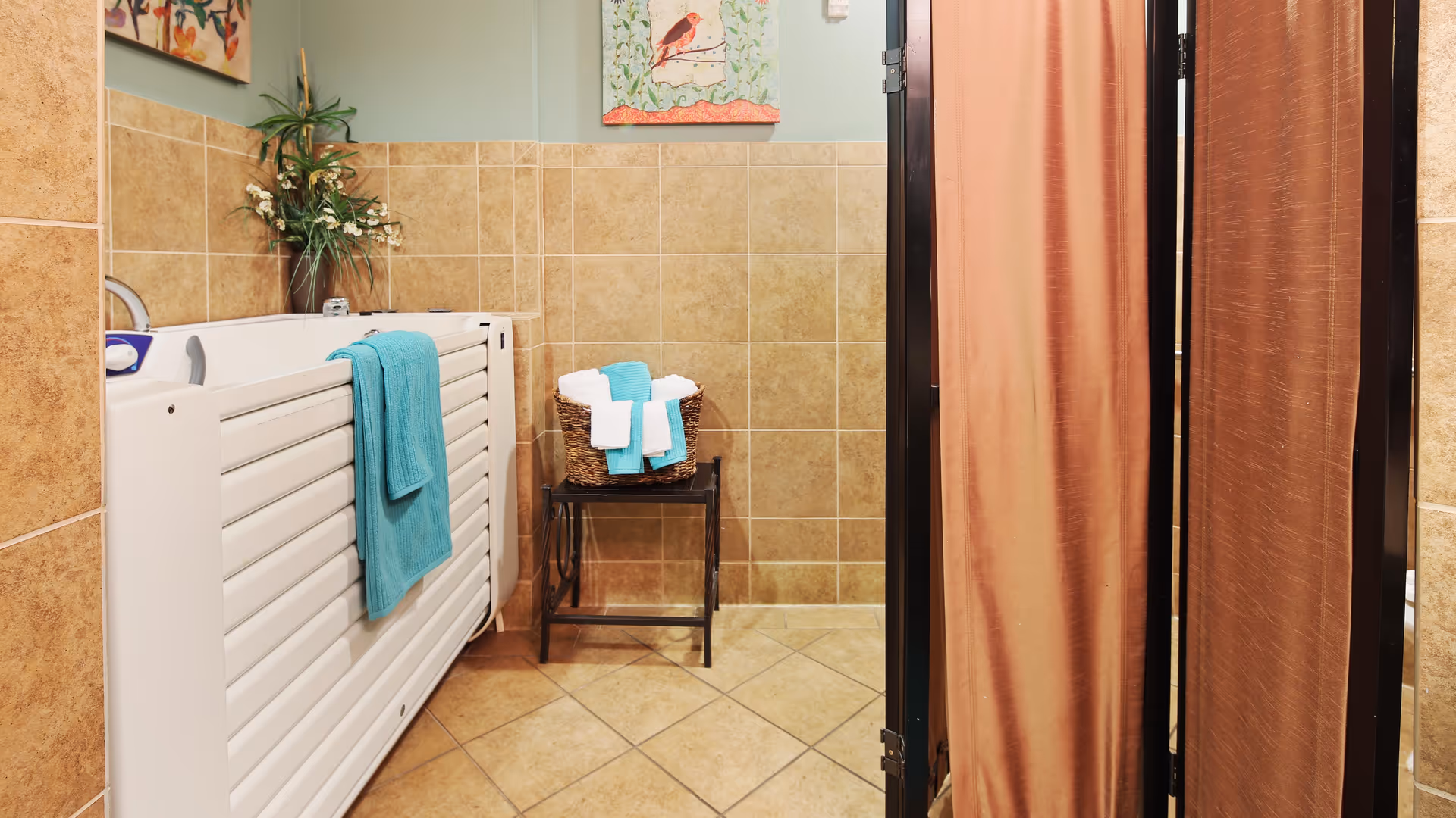 A tiled room with a white walk-in bathtub on the left side, a turquoise towel hanging on the tub, a small table with a basket holding rolled white and turquoise towels, a folding privacy screen on the right, and two framed bird paintings on the wall above the tub.
