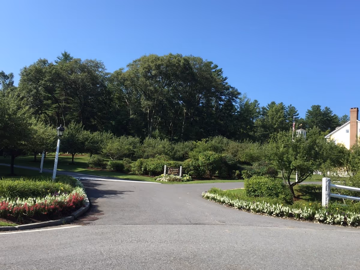 A paved driveway splits into two paths surrounded by well-maintained greenery and flower beds with white and red flowers. There are trees and bushes on both sides, and a white fence is visible on the right. In the background, there is a yellow building with a chimney and an American flag. The sky is clear and blue.