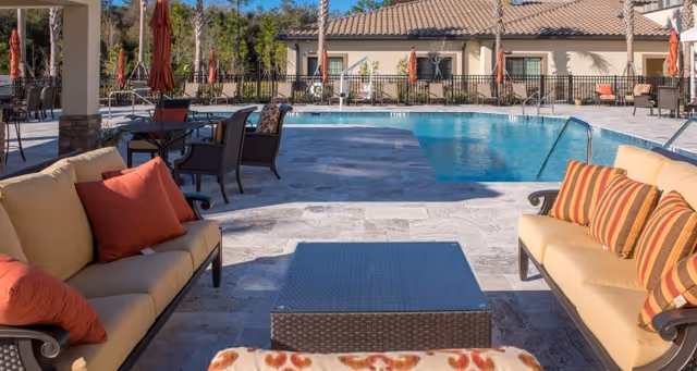 Outdoor seating area with cushioned sofas and chairs around a rectangular coffee table next to a swimming pool. The pool area is surrounded by a fence, with a building and trees in the background. Several umbrellas are closed and positioned around the pool.
