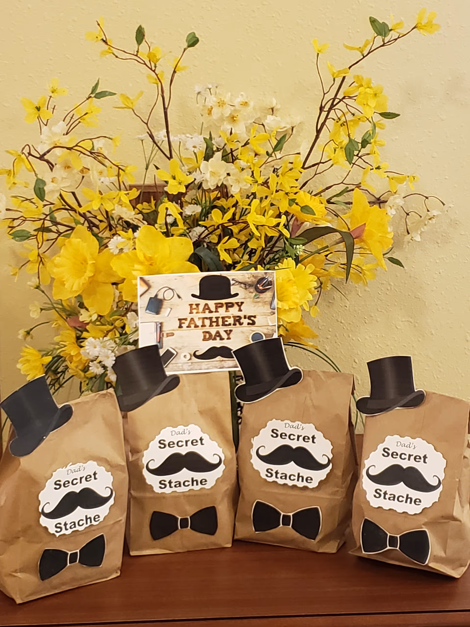 Four Father's Day gift bags decorated with mustaches, top hats, and bow ties arranged in front of a large yellow floral display.