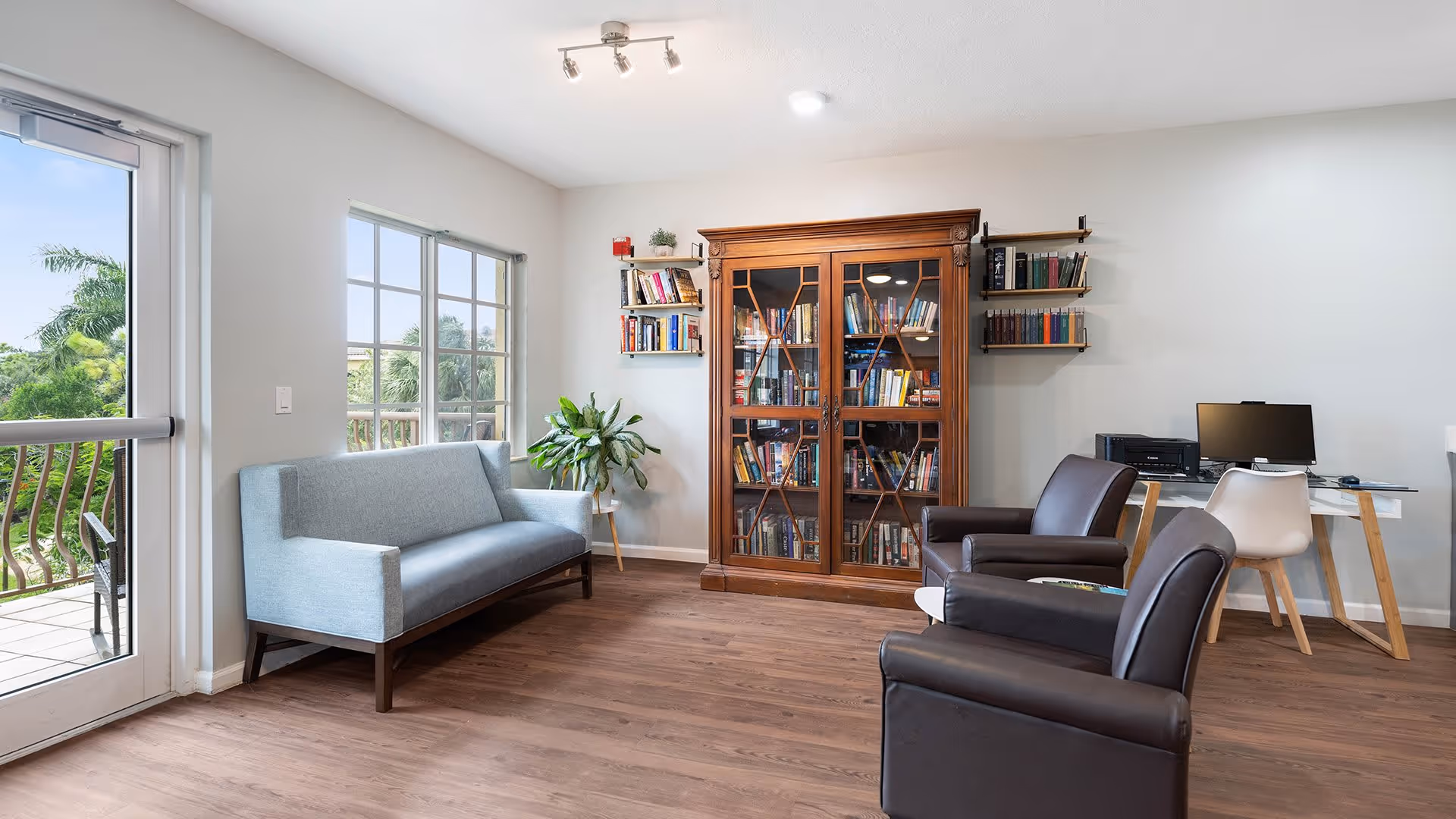 A bright and cozy living room area with a light blue sofa near a glass door leading to a balcony with greenery outside. There is a wooden bookcase filled with books against the wall, flanked by two small wall-mounted bookshelves. Two dark brown armchairs face a small white table. In the corner, there is a desk with a computer and printer, accompanied by a white chair. The room has wooden flooring and white walls.