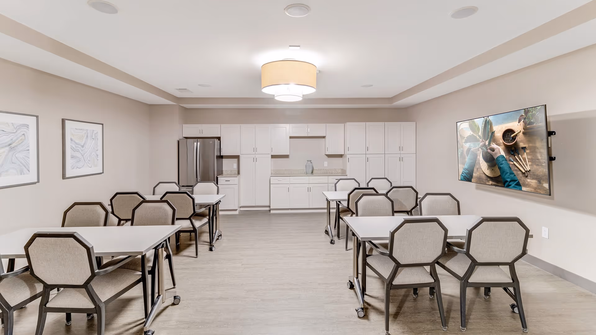 A modern dining room with several tables and chairs arranged neatly. The room features light-colored walls, a large ceiling light fixture, framed artwork on one wall, and a flat-screen TV mounted on the opposite wall. At the far end, there is a kitchenette area with white cabinets, a refrigerator, and a countertop.