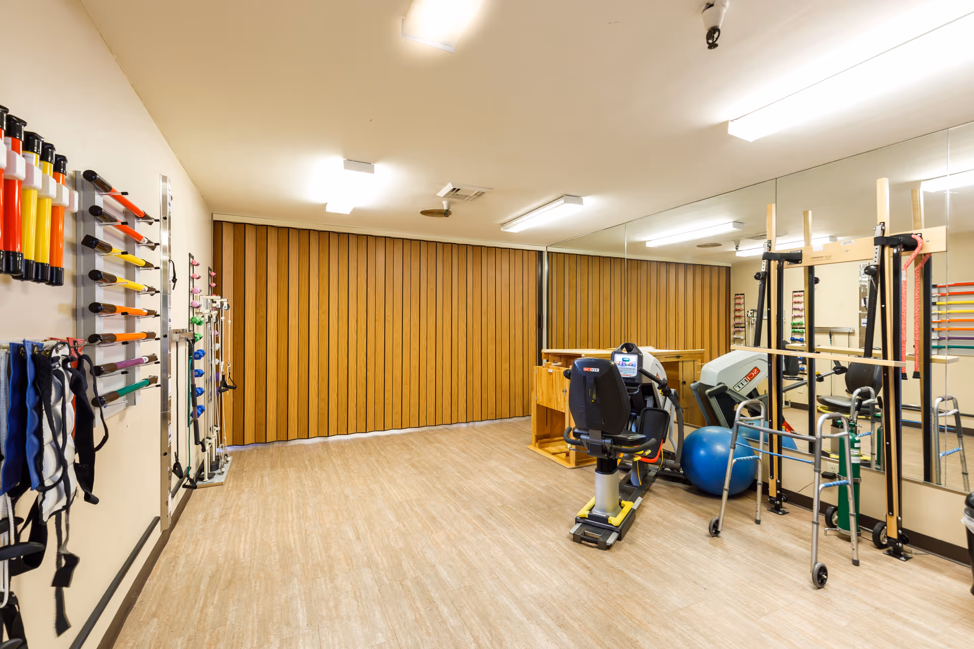 A well-lit physical therapy or exercise room with various rehabilitation equipment including resistance bands, weights, a stationary exercise bike, a walker, a blue exercise ball, and a wooden balance or stretching frame. The room has wood paneling on one wall, large mirrors on another, and light-colored flooring.